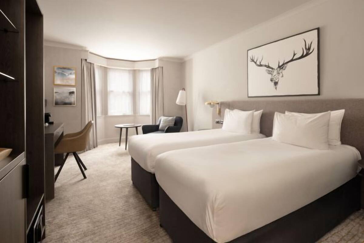 A hotel room with two beds, a desk, a chair, a small table, and a window with curtains.