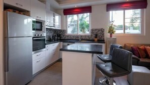 A modern kitchen with stainless steel appliances, a central island, and a dining area with a black chair. - Orchid Paradise Homes OPV 213 (Hua Hin)