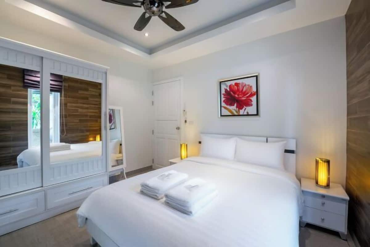 Une chambre à coucher moderne avec un grand lit, des lampes de chevet, un ventilateur de plafond et une œuvre d’art florale encadrée accrochée au mur.