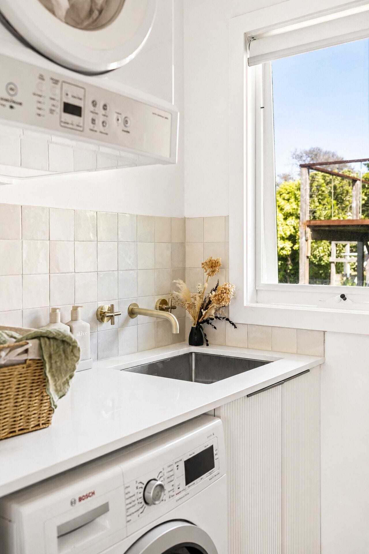 A modern laundry room with a Bosch washing machine, a window with a view of greenery, and a white tiled backsplash.