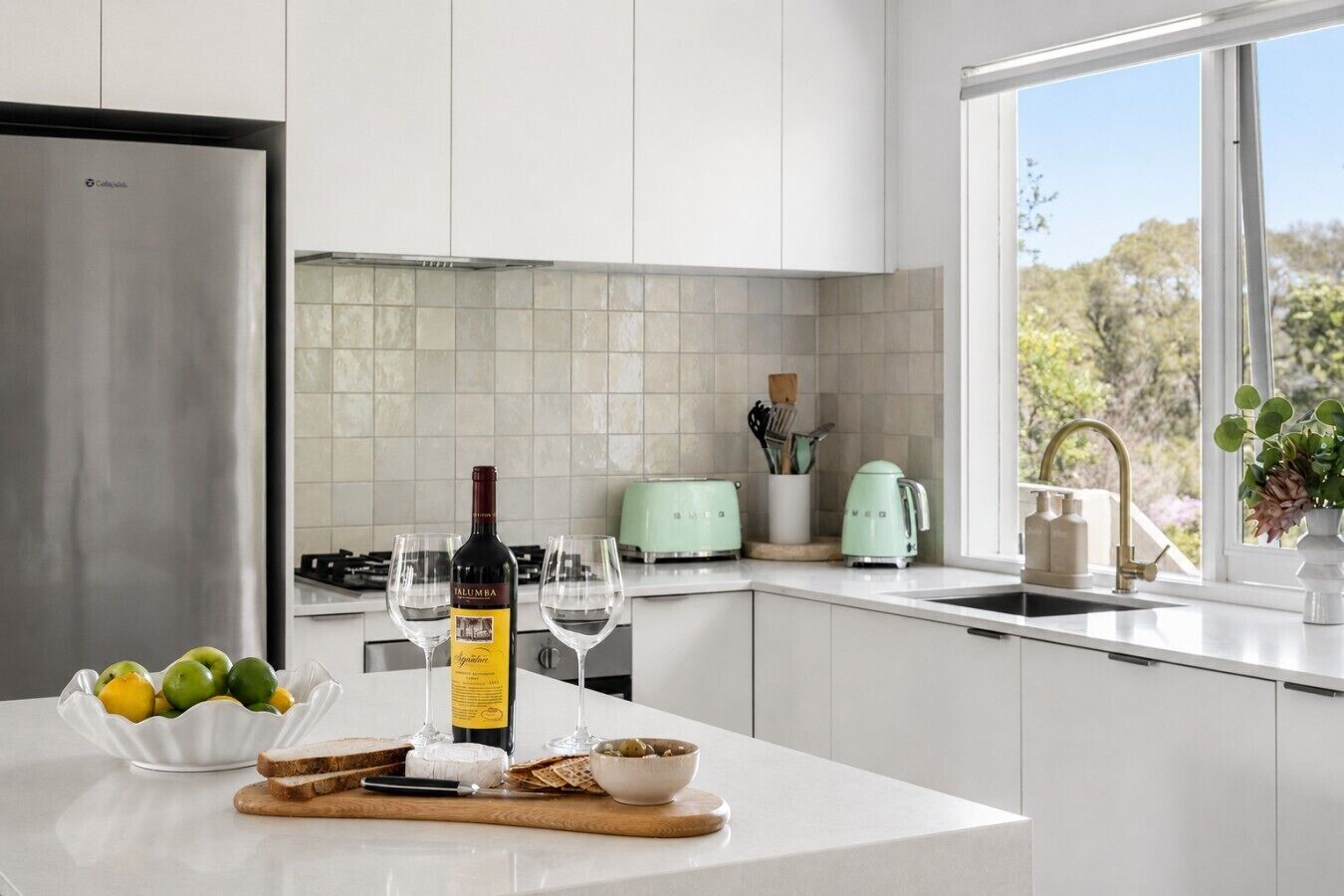 A modern kitchen with a stainless steel refrigerator, a wine bottle, glasses, and a bowl of fruit on the counter.