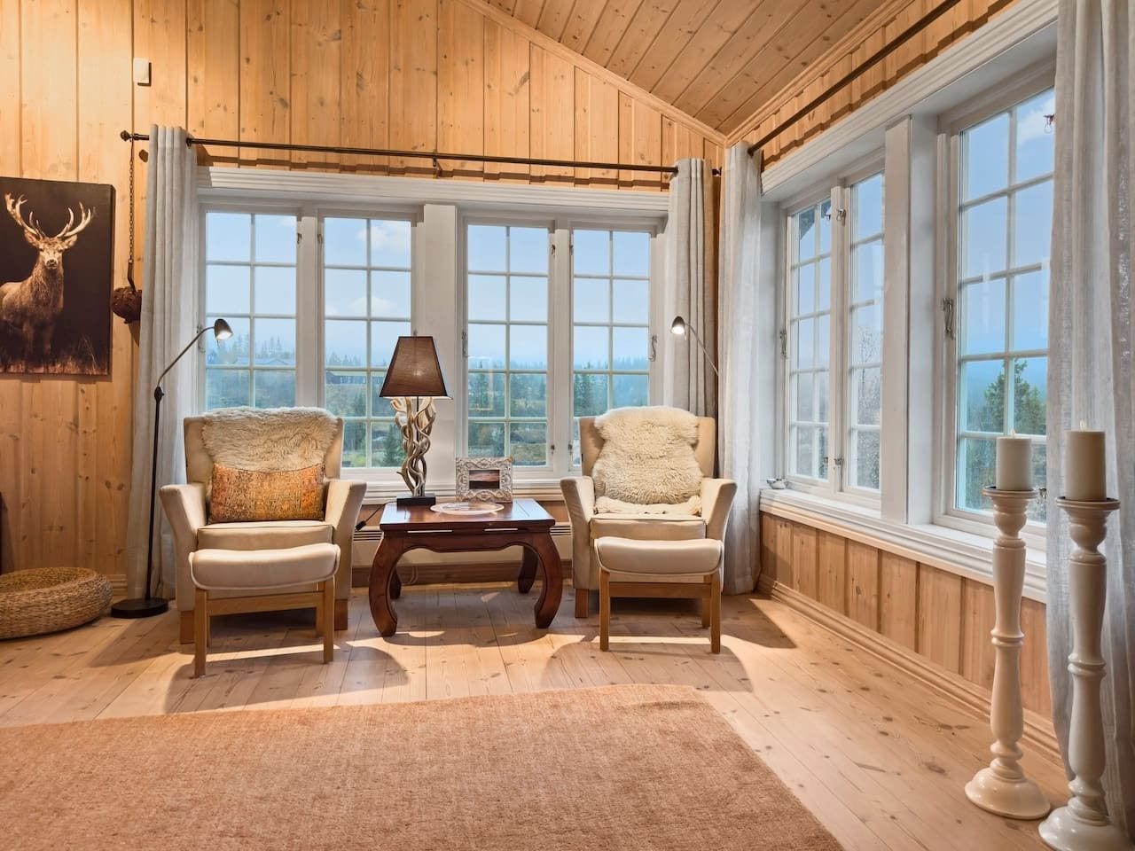 A room with wooden paneled walls, two chairs with fur throw pillows, a table with a lamp, and large windows with curtains.