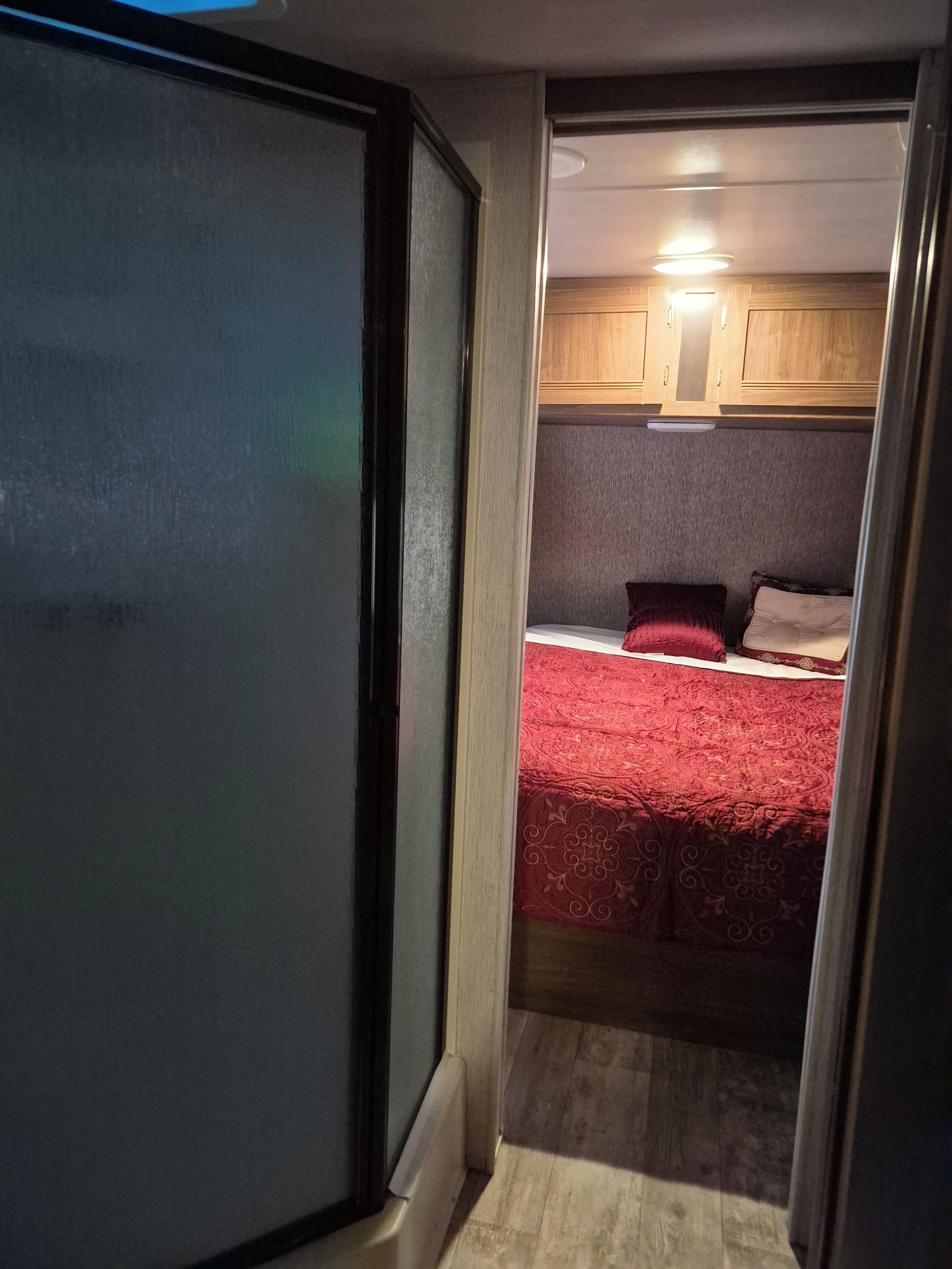 Ein schmaler Flur, der in ein Schlafzimmer mit roter Bettdecke, Holzschränken und einer Deckenleuchte führt.