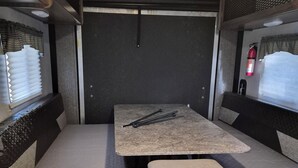 Interior compacto de una autocaravana con una mesa de comedor, cuatro asientos acolchados, una pequeña cocina y una ventana con persianas.