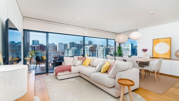 A modern living room with a large window, a sofa, a dining area, and a city view. - Miraflores Modern Apt w/ Balcony by LovelyStay (Algés)