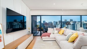 A modern living room with a large TV, a sofa with cushions, a coffee table, and a city view through large windows. - Miraflores Modern Apt w/ Balcony by LovelyStay (Algés)
