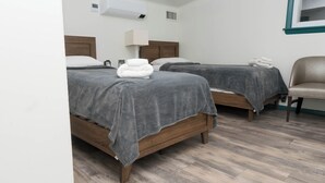 Une chambre d’hôtel avec deux lits en bois, des couvre-lits gris, des serviettes blanches, un plancher en bois et une fenêtre encadrée de bleu sarcelle.