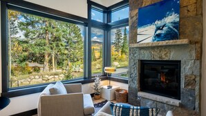 Una sala de estar moderna con chimenea de piedra, ventanas amplias que ofrecen vista a un bosque y un área de estar acogedora.