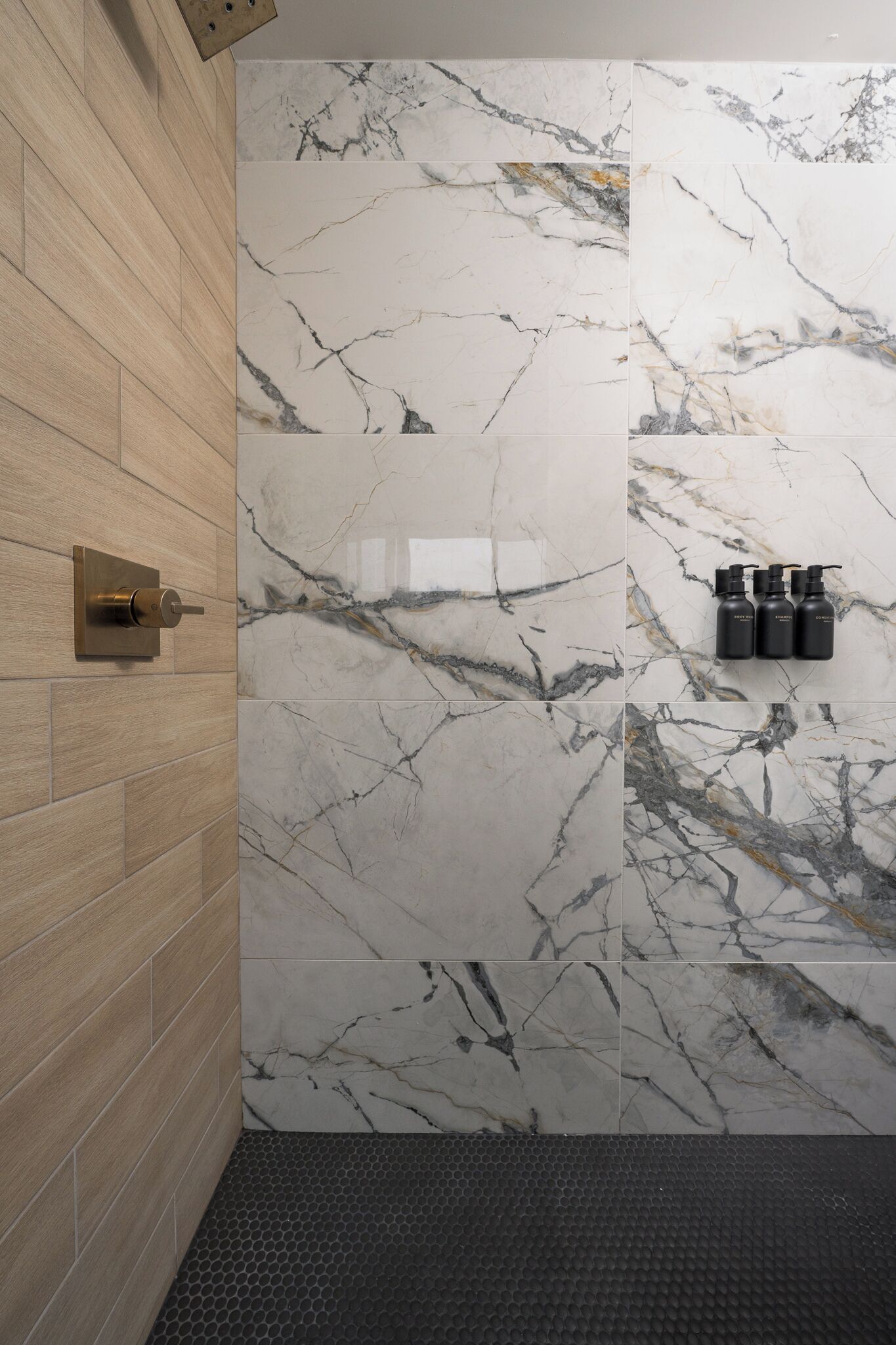 Une salle de bain avec des carreaux de marbre, un mur en bois, un sol noir texturé et une porte dotée d’une poignée moderne.