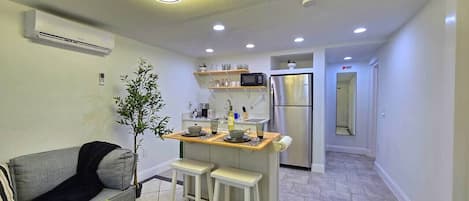 Un espace de vie compact comprenant un coin cuisine, un coin repas avec des tabourets, un canapé gris et une petite plante en pot.