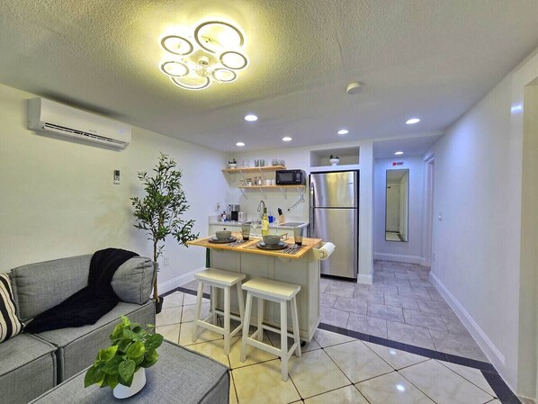 Un espacio habitable compacto con kitchenette, una zona de comedor con taburetes, un sofá gris y una pequeña planta en maceta.