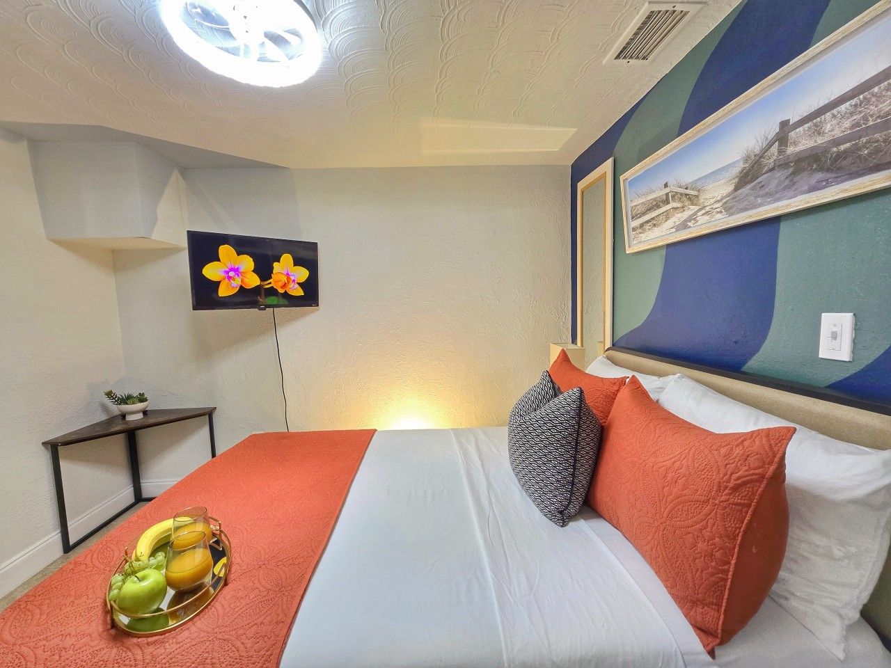 A hotel room with a bed, a flat-screen TV, a wall-mounted picture, and a small table with a fruit tray.