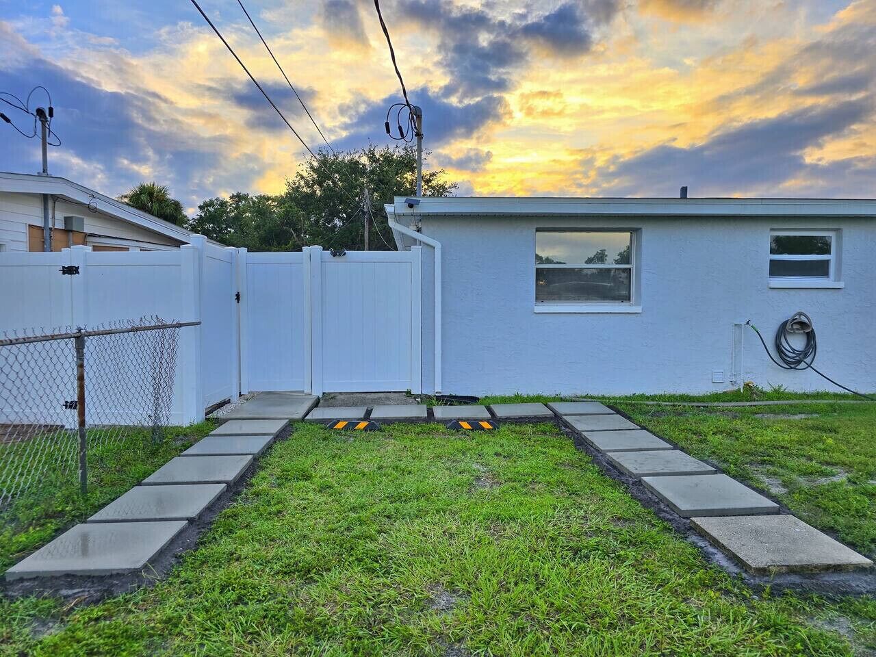 A residential backyard with a concrete pathway leading to a white house, surrounded by a chain-link fence.