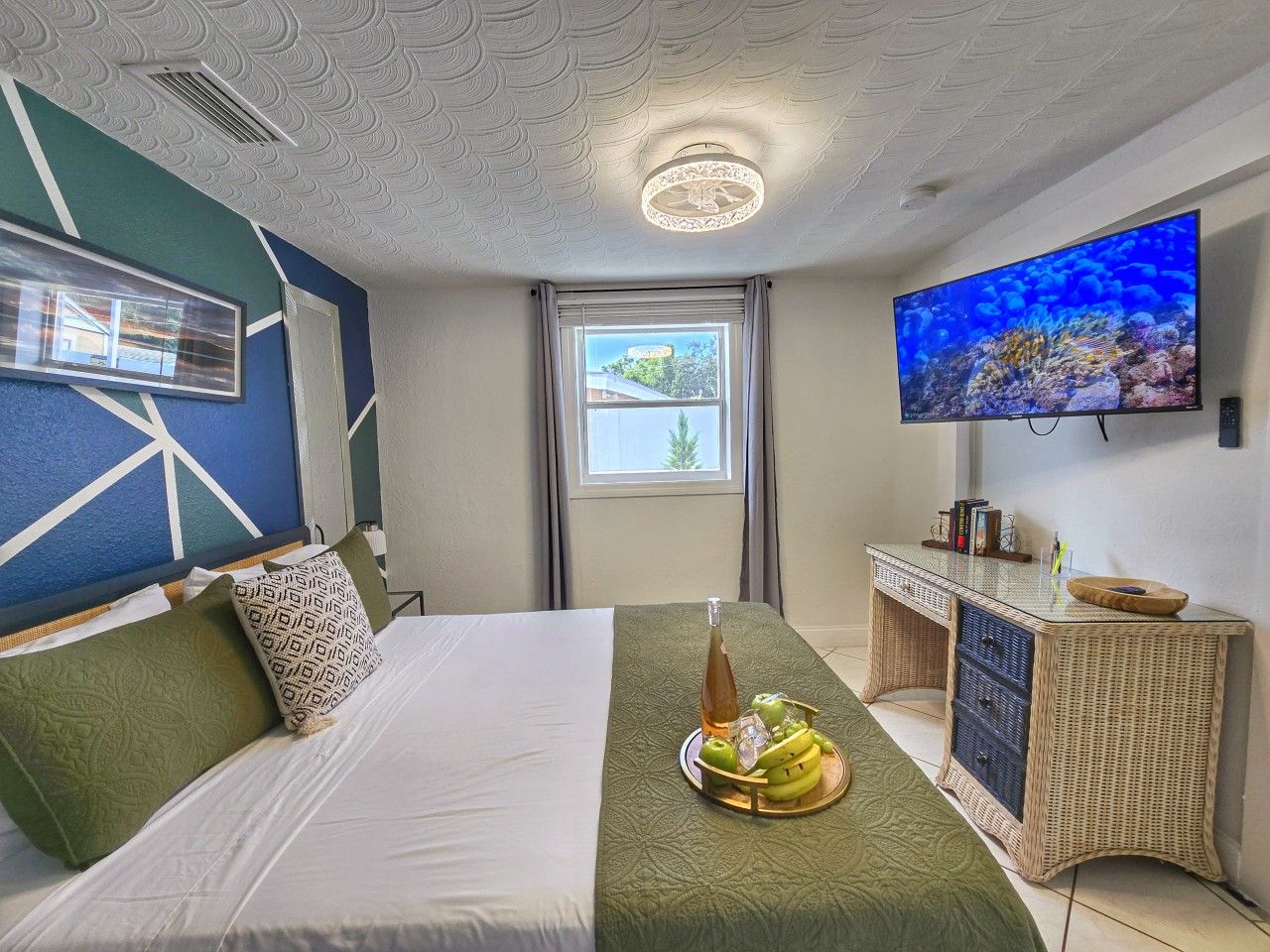 A hotel room with a bed, a television, a window, and a wicker side table with a tray of fruit.