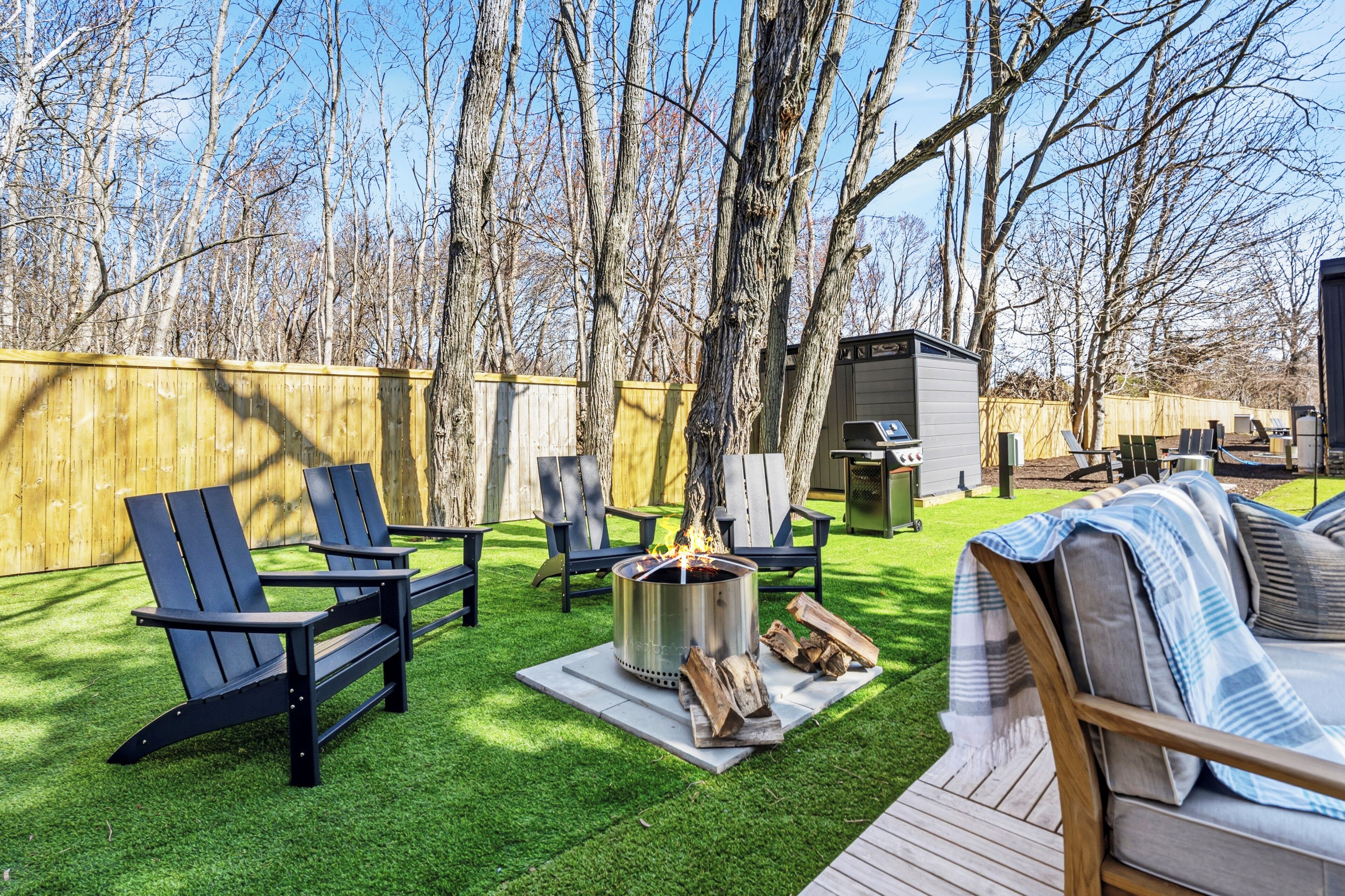 Un patio trasero con un hoyo de fuego, sillas Adirondack, una terraza de madera y una zona cubierta para la parrilla.