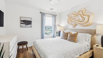 Un dormitorio con cama, mesita de noche, lámpara, ventana con cortinas, una obra de arte en la pared y una planta.