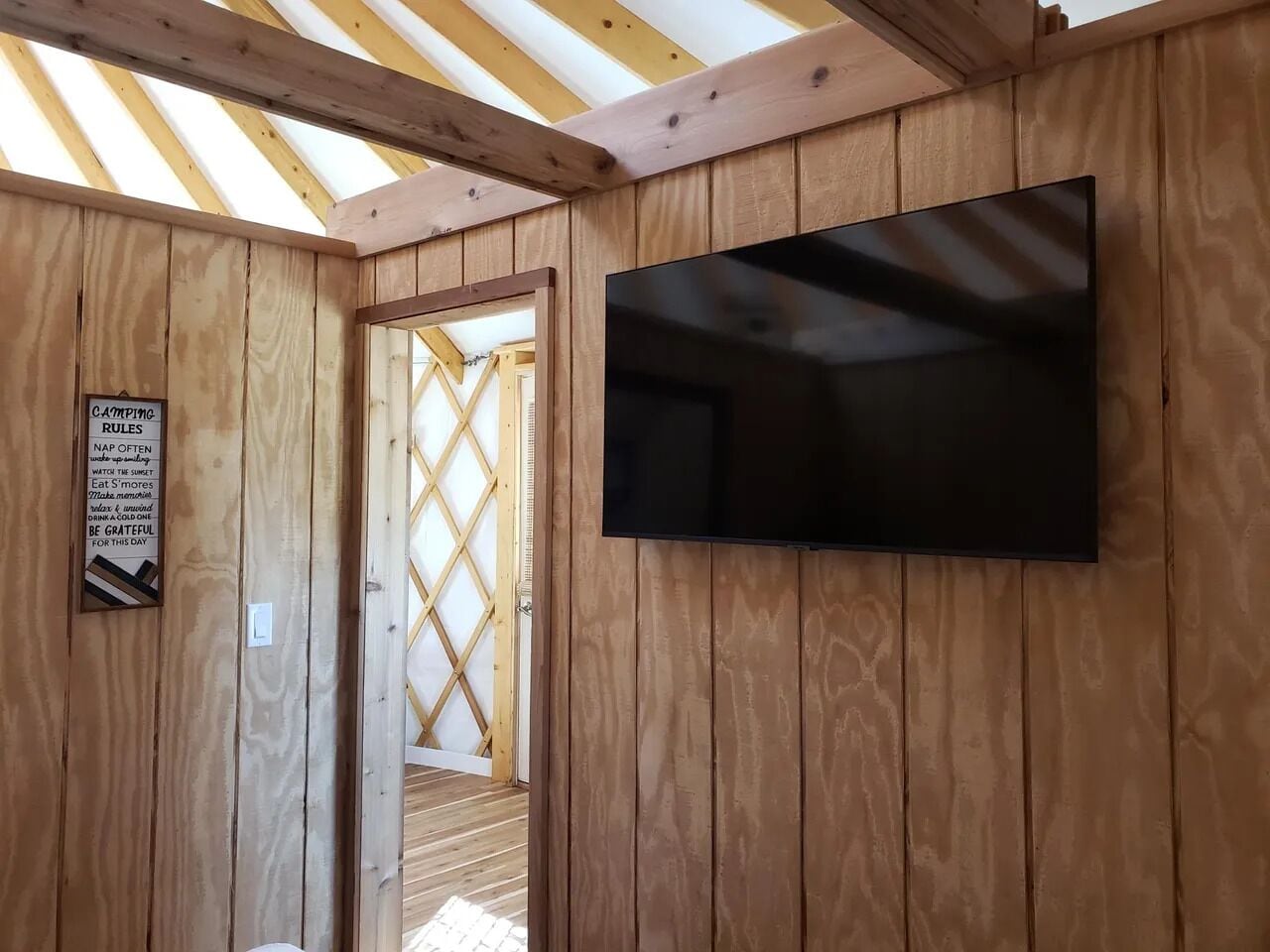 A wooden cabin with a TV mounted on the wall, a door leading to an outdoor area, and a sign with camping rules.