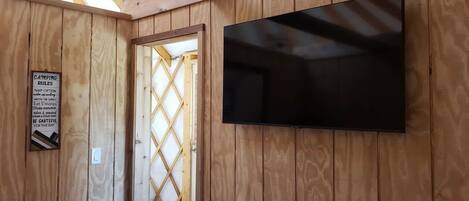 A wooden cabin with a TV mounted on the wall, a door leading to an outdoor area, and a sign with camping rules.
