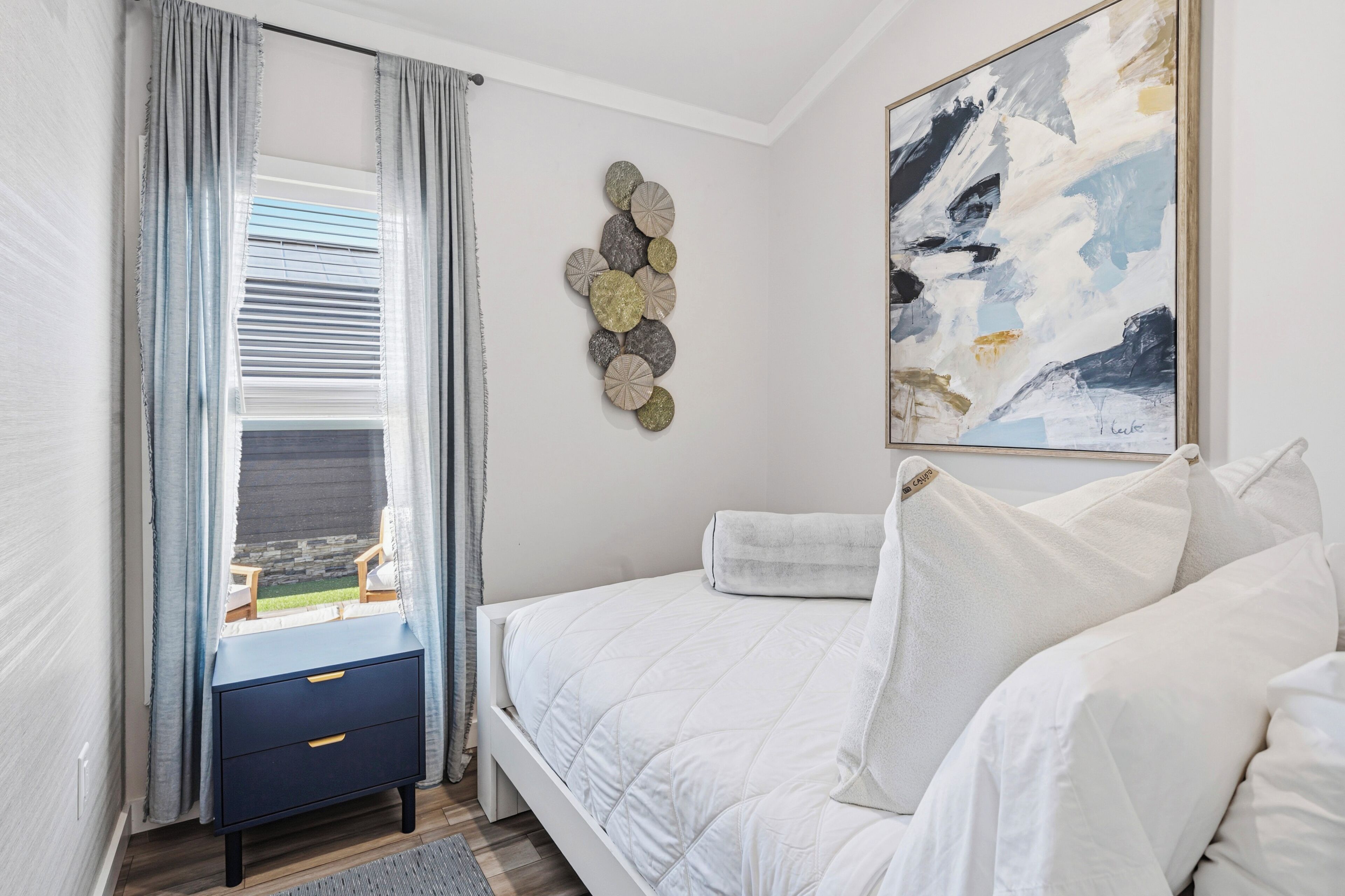 A bedroom with a bed, a nightstand, a window with curtains, a wall art piece, and a view of the outdoors.