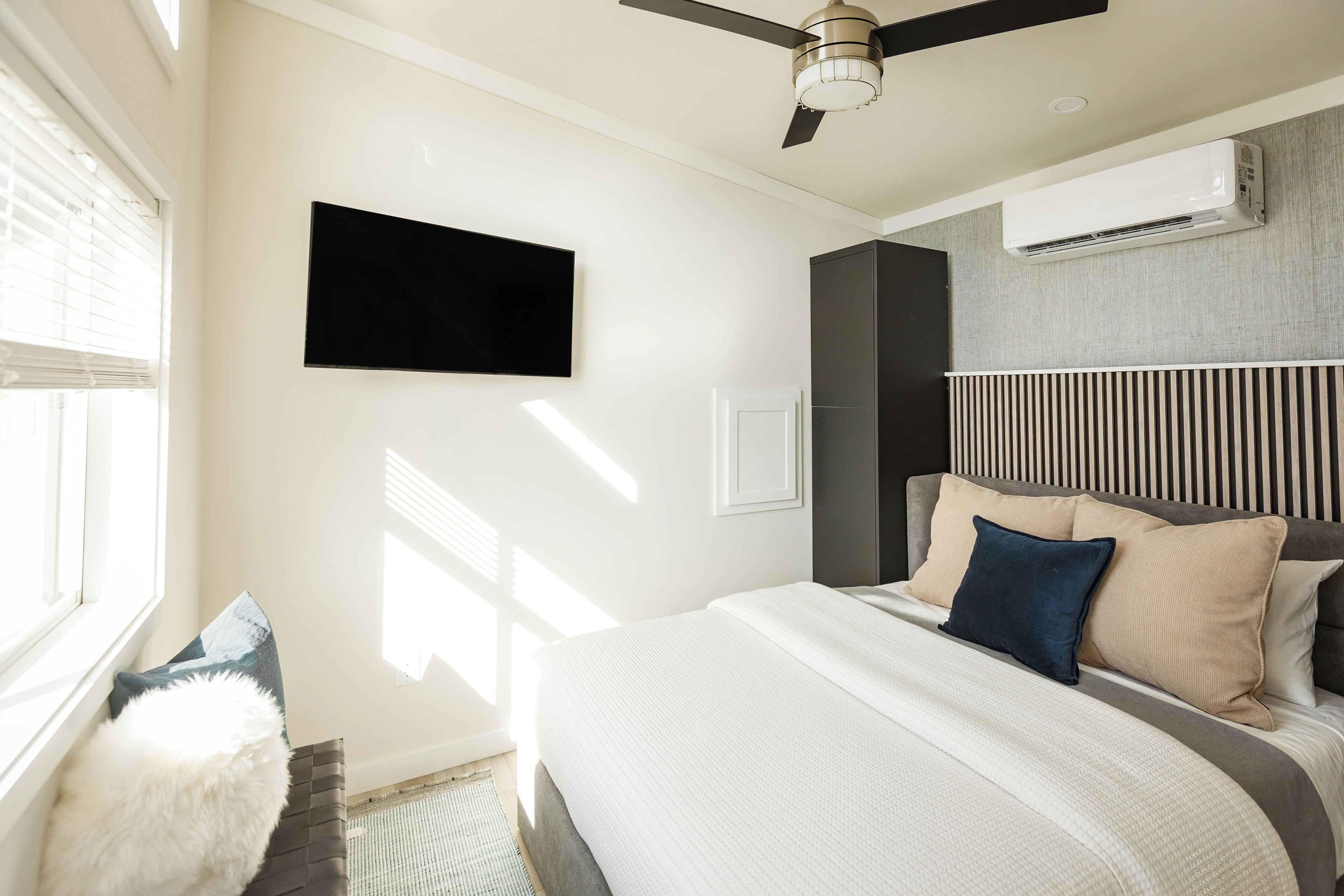 A modern bedroom with a bed, a ceiling fan, a mounted TV, and an air conditioning unit.