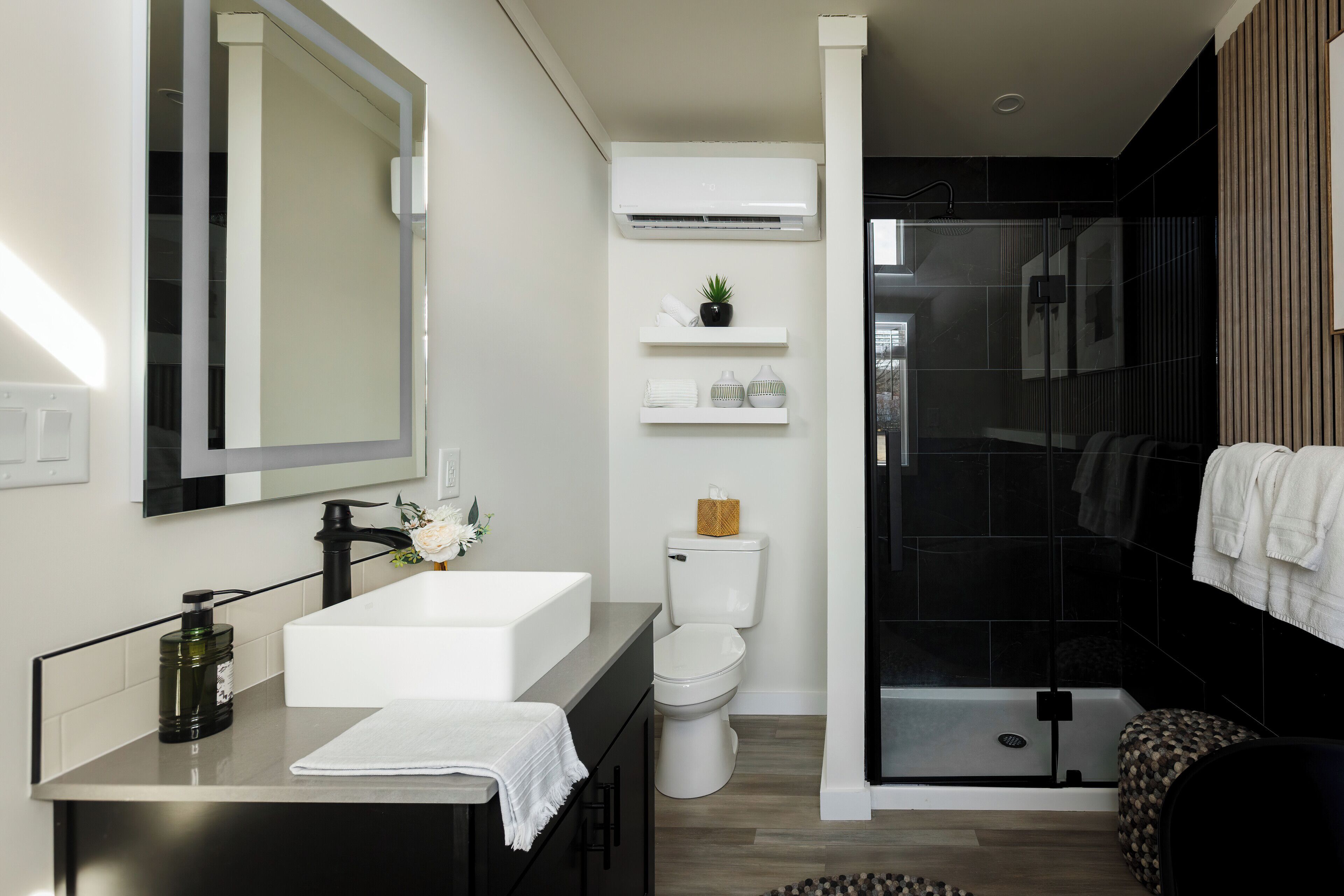 A modern bathroom with a white sink, black faucet, and a large mirror. There is a toilet with a basket on the tank and a shelf above it. A glass shower enclosure is visible to the right.