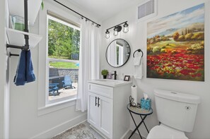 Et badeværelse med et hvidt toiletbord, et rundt spejl, et toilet og et vindue med udsigt til en have.