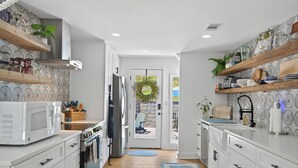 A modern kitchen with white cabinetry, a microwave, oven, and a sink. There are wooden shelves with plants and kitchenware. A door leads to an outdoor area with a chair and a wreath. - Lake house views, w/dock in quiet cove, new renovations & great outdoors spaces! (Dahlonega)