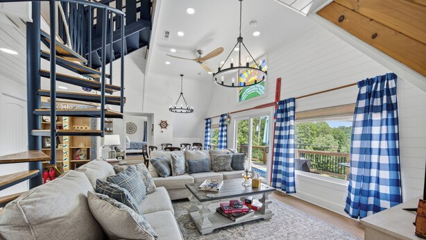 A spacious living room with a high ceiling, a staircase, a large sofa, a coffee table, and a view of the outdoors. - Lake house views, w/dock in quiet cove, new renovations & great outdoors spaces! (Dahlonega)