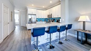 Een moderne keuken met witte kastjes, een centraal kookeiland met barstoelen en een eethoek met een tafel en lamp.