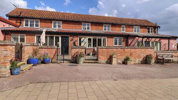 A two-story brick building with a tiled roof, a front entrance with a black door, and a small porch area with potted plants. - Coachman's Cottage (Ipswich)