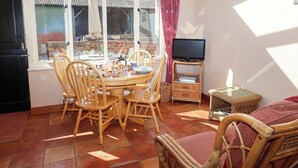A dining area with a table set for four, surrounded by wicker chairs, a television on a stand, and a view of a brick building through large windows. - Coachman's Cottage (Ipswich)