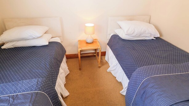 A room with two beds, each with white pillows and blue bedding with white polka dots. There is a small wooden nightstand with a lamp between the beds. - Churn House (Ipswich)