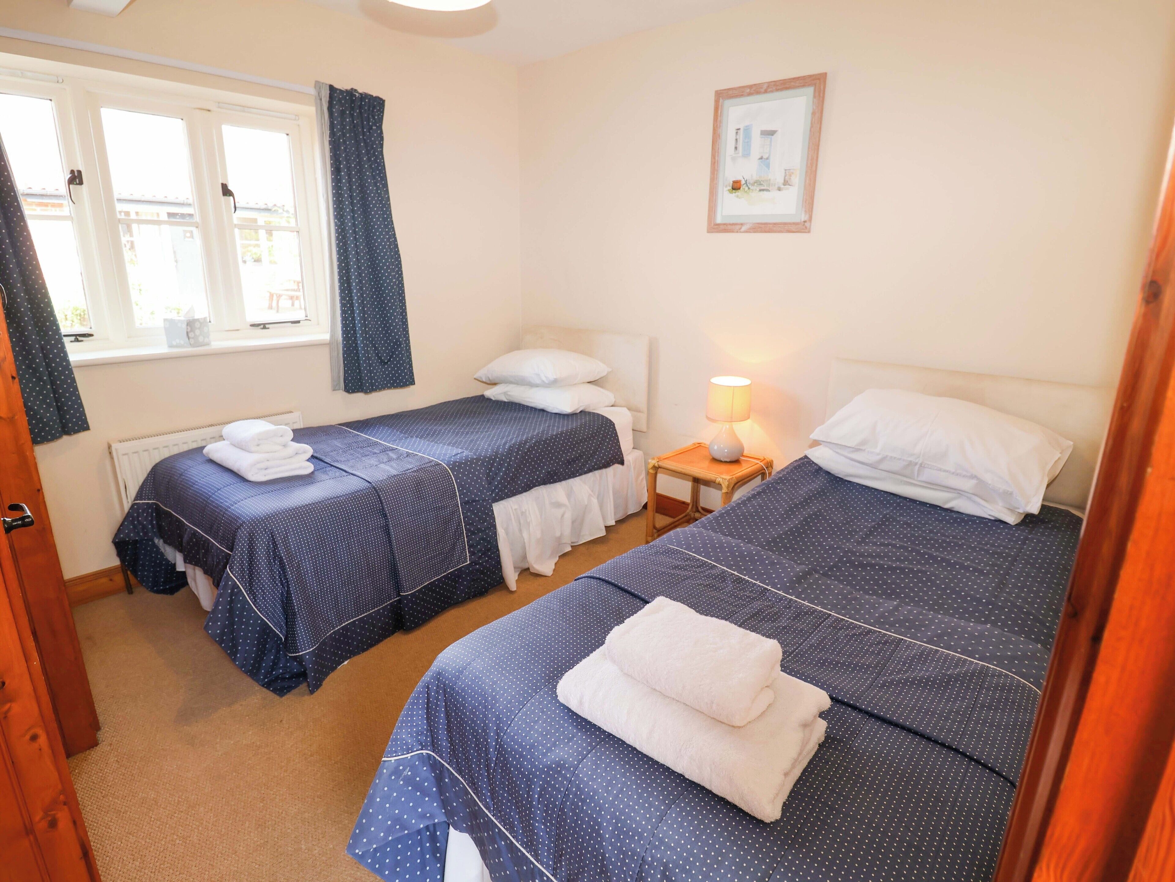 A hotel room with two single beds, a bedside table with a lamp, a window with curtains, and a framed picture on the wall.