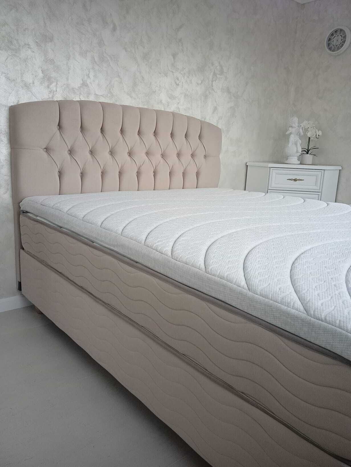 Een bed met een gestoffeerd hoofdeinde, een wit matras en een nachtkastje met een kleine plant.