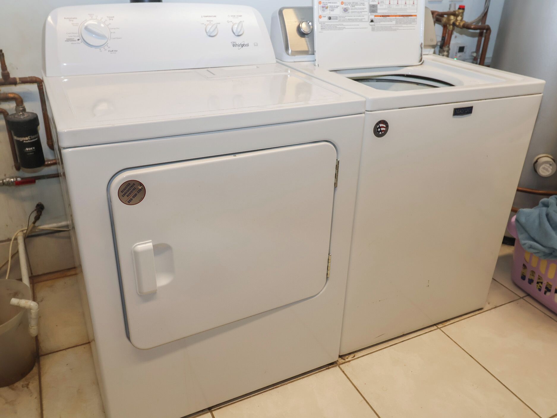Lavadora y secadora Whirlpool blancas de carga frontal en un cuarto de lavado, con tuberías visibles y una cesta de ropa rosa.
