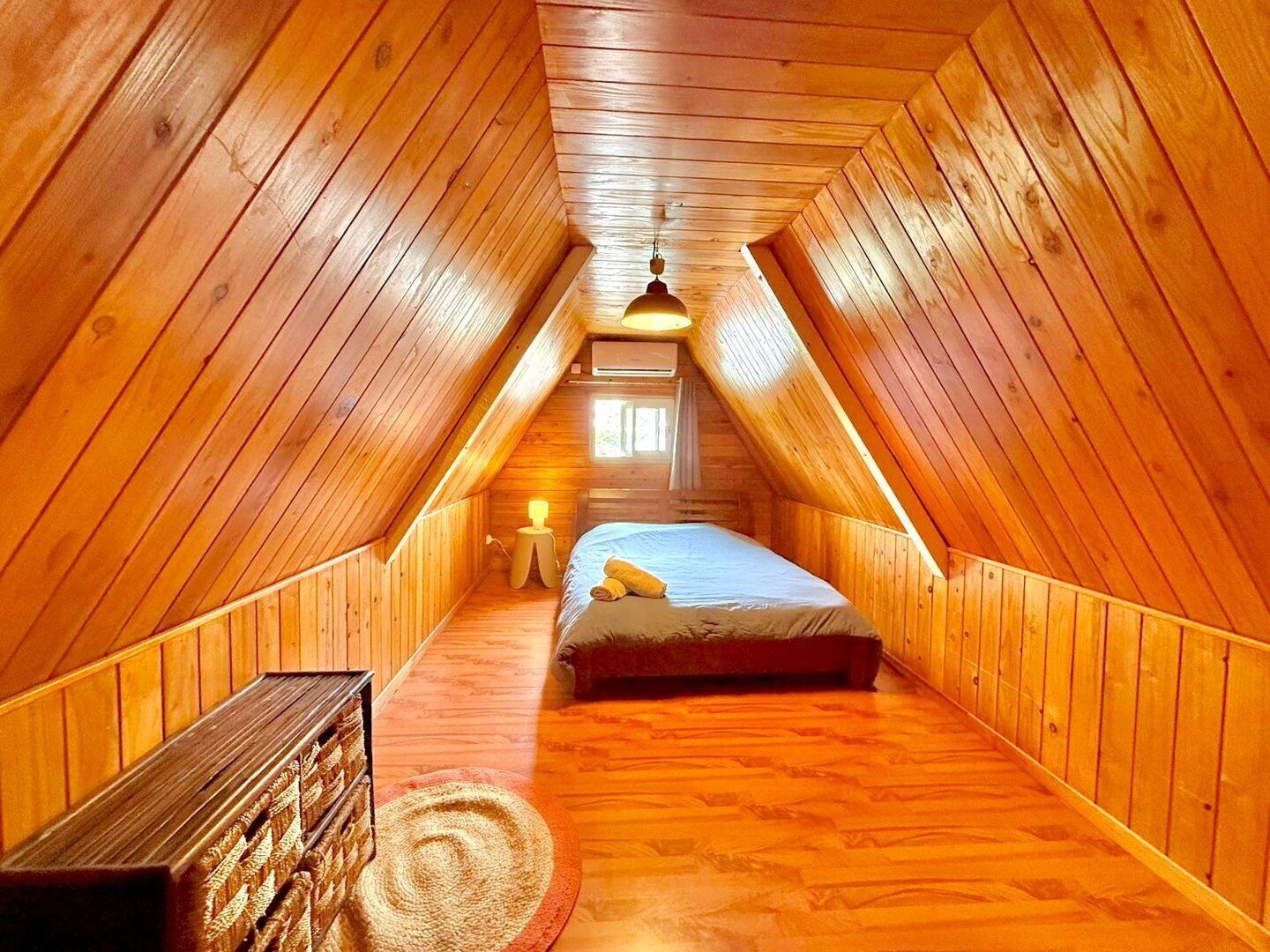 A cozy attic bedroom with wooden walls and a sloped ceiling, featuring a bed, a small table, and a window.