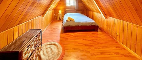A cozy attic bedroom with wooden walls and a sloped ceiling, featuring a bed, a small table, and a window.