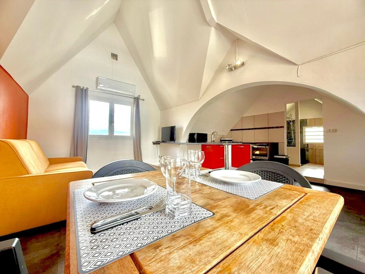 Comedor con mesa de madera preparada para cuatro personas, sofá amarillo y cocina al fondo.
