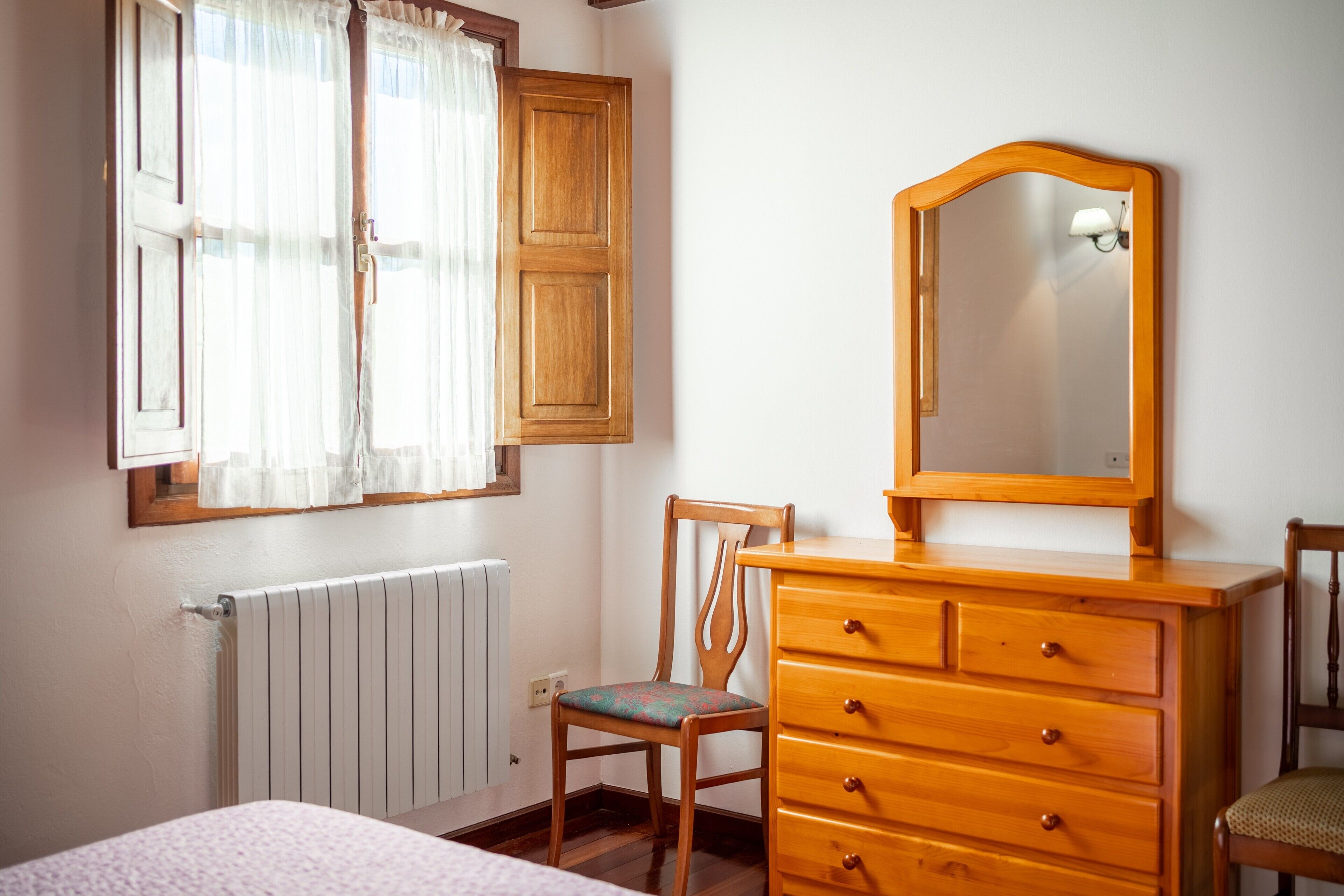 A bedroom with a wooden dresser, a chair, a bed, a window with curtains, and a radiator.