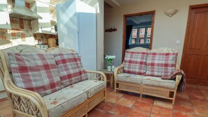 A room with two wicker sofas with plaid cushions, a tiled floor, a wooden door, and a kitchen area in the background. - Wheelwright's Cottage (Ipswich)