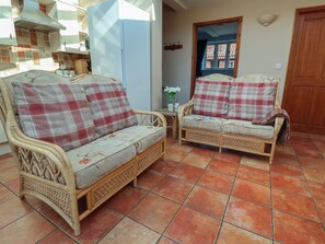 A room with two wicker sofas with plaid cushions, a tiled floor, a wooden door, and a kitchen area in the background. - Wheelwright's Cottage (Ipswich)