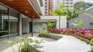A modern building with a courtyard featuring plants, a bench, and a curved seating area. - Aptos modernos em prédio com piscina a 300m do metrô - UOYs (São Paulo)