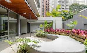 A modern building with a courtyard featuring plants, a bench, and a curved seating area. - Aptos modernos em prédio com piscina a 300m do metrô - UOYs (São Paulo)