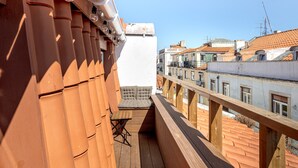 A balcony with a wooden floor, a cushioned seating area, and a view of neighboring buildings.