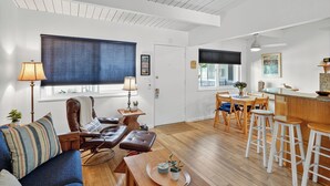 A modern living room with a sofa, armchair, and coffee table. There is a dining area with a table and chairs, and a kitchen area with a bar counter and stools. - NEW! 1-bed condo in Carp, Steps to Sand! (Carpinteria)