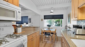 A modern kitchen with wooden cabinets, a granite countertop, a built-in microwave, a gas stove, and a dining area with a table and chairs. - NEW! 1-bed condo in Carp, Steps to Sand! (Carpinteria)
