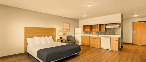 A hotel room with a bed, a desk, a chair, a kitchenette, and a door.