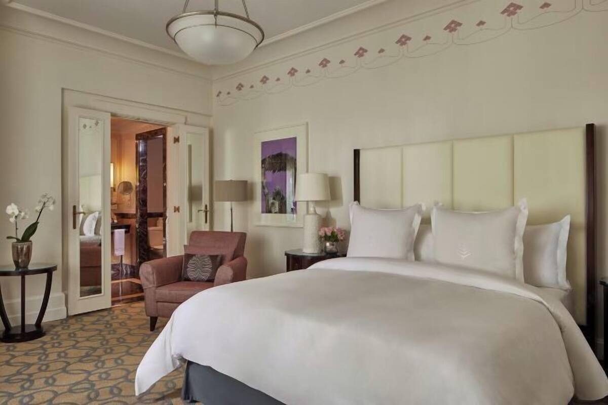 Habitación de hotel con una cama grande, una silla, una mesita de noche, una lámpara y una puerta que da a otra habitación.