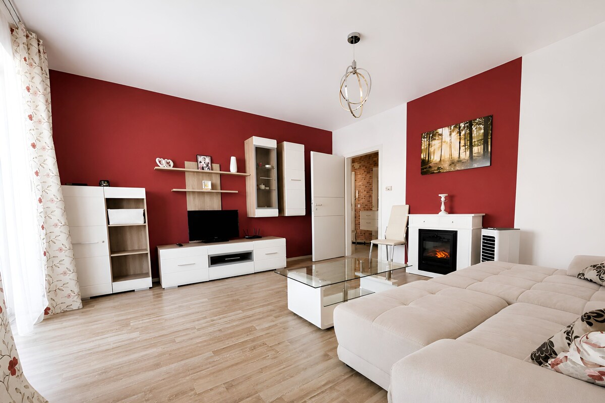 A modern living room with a red accent wall, a glass coffee table, a white sofa, and a fireplace.