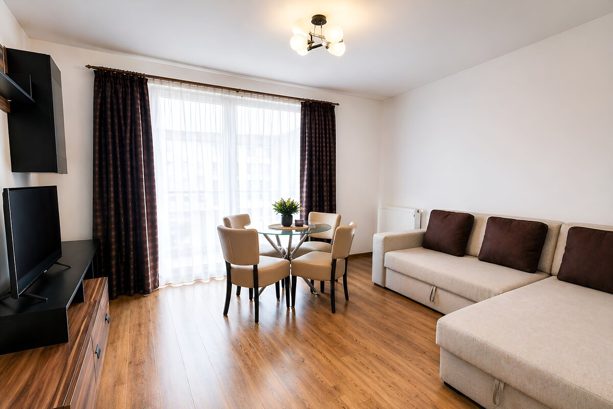 A modern living room with a flat-screen TV, a sofa, a dining table with chairs, and a potted plant.